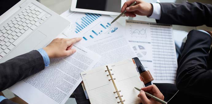 Overhead view of hands pointing at charts and documents on a desk with a laptop and tablet, blue and white tones, professional mood