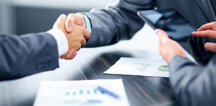 Close-up of two people in suits shaking hands over a desk with charts and a tablet, gray and blue tones, professional mood